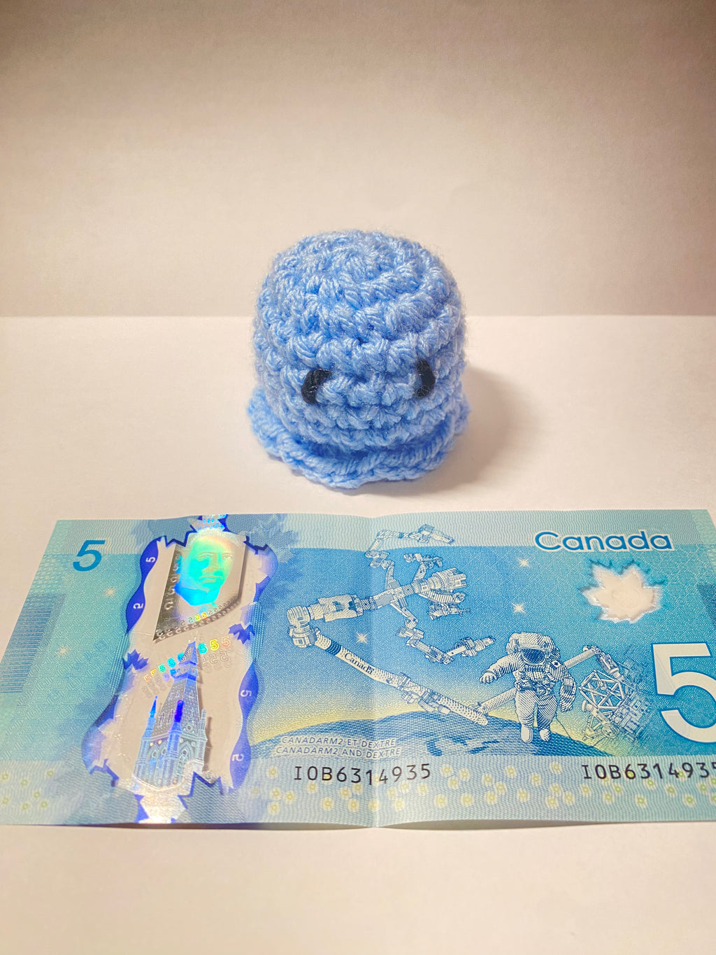 Blue crocheted toy next to a Canadian five-dollar bill on a white surface