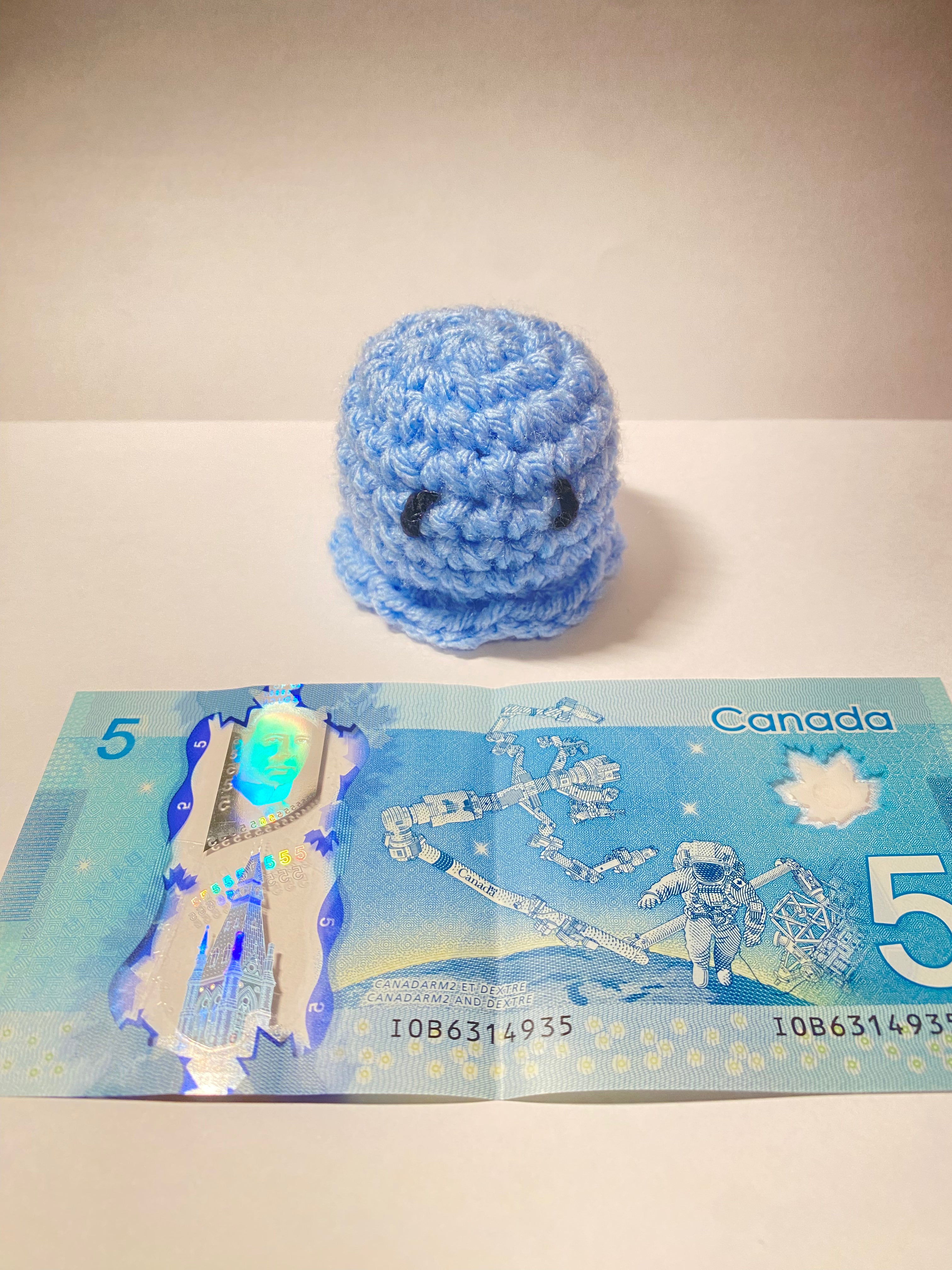 Blue crocheted toy next to a Canadian five-dollar bill on a white surface