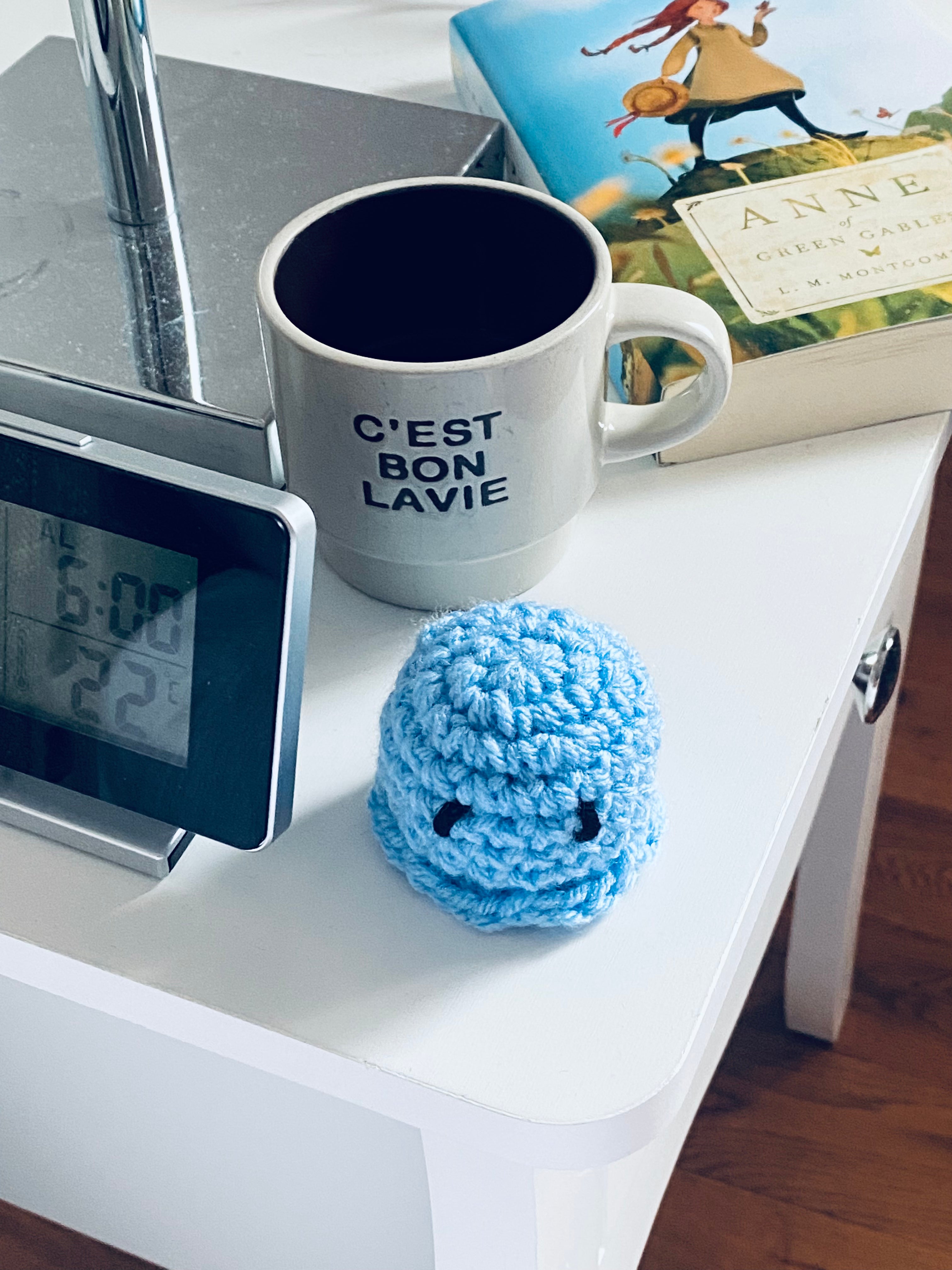White mug with 'C'EST BON LAVIE' text, blue crocheted octopus, digital clock, and book on a white surface.
